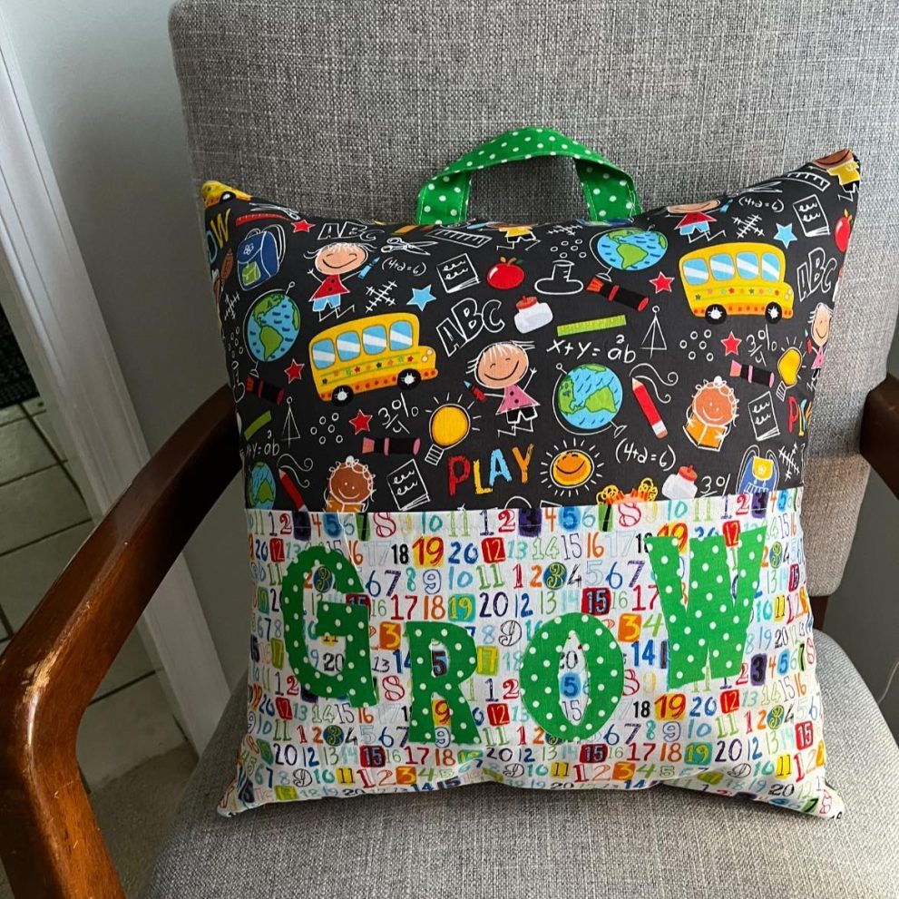 A handmade children's reading pillow with a colorful school-themed fabric, featuring playful designs of school buses, numbers, and doodles. The front pocket displays the word "GROW" in green polka-dot letters, and a green handle is attached to the top. Made by Stitches by Sharon Lane of the Lehigh Valley PA